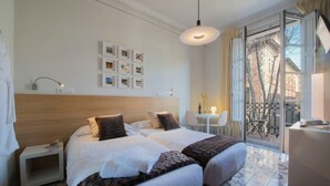 Ein Hotelzimmer mit zwei Betten, einem kleinen Schreibtisch, einem Stuhl, einem Tisch mit einer Lampe und einem Balkon mit Geländer.