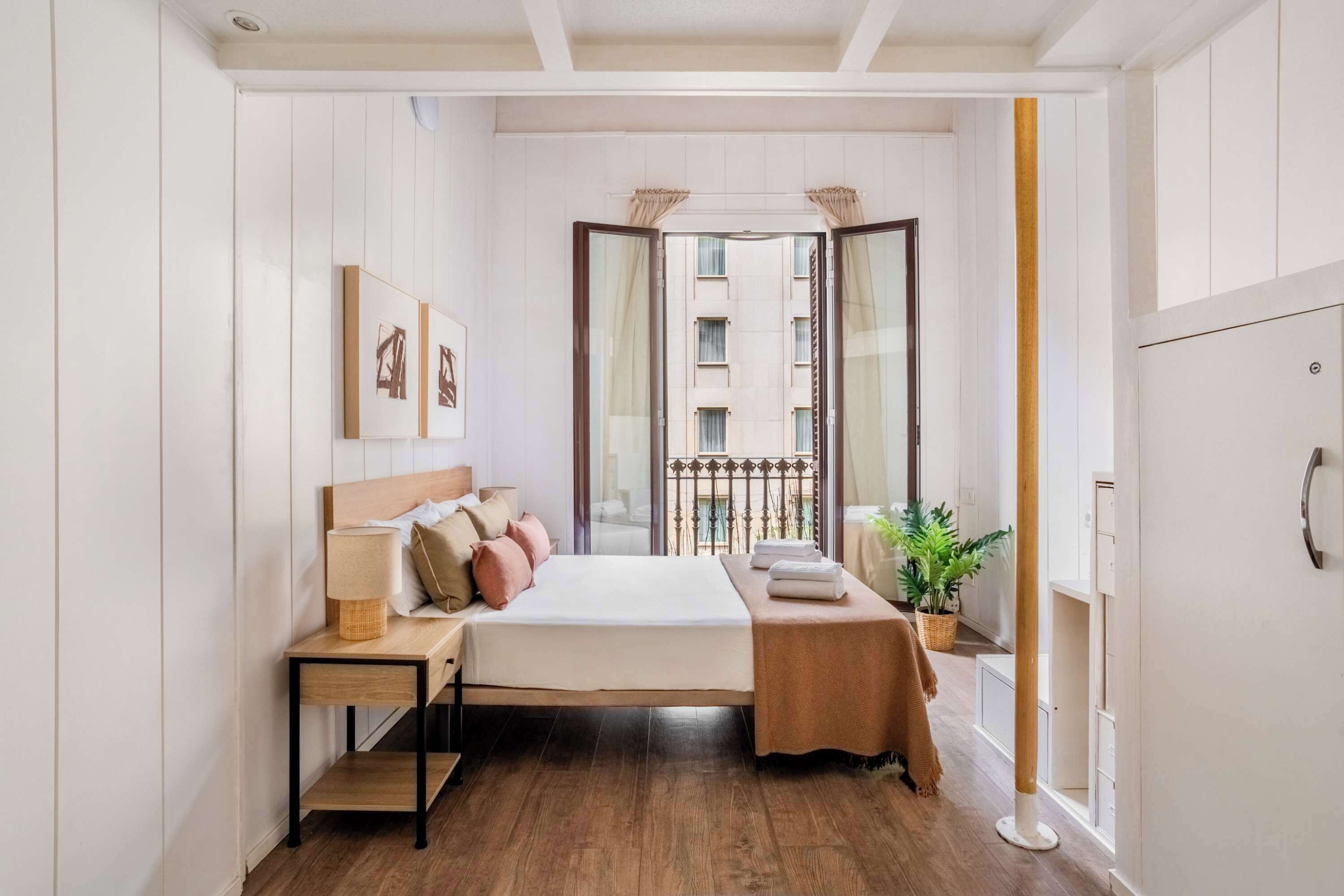 Quarto com cama, mesa de cabeceira, luminária e vista para um edifício através da janela.