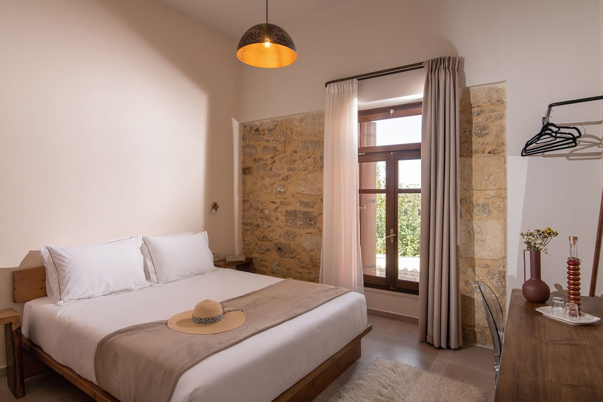 Un dormitorio con pared de piedra, una cama con ropa de cama blanca, una mesita de noche de madera, una lámpara colgante, una ventana con cortinas y un aparador de madera con un florero y una botella.