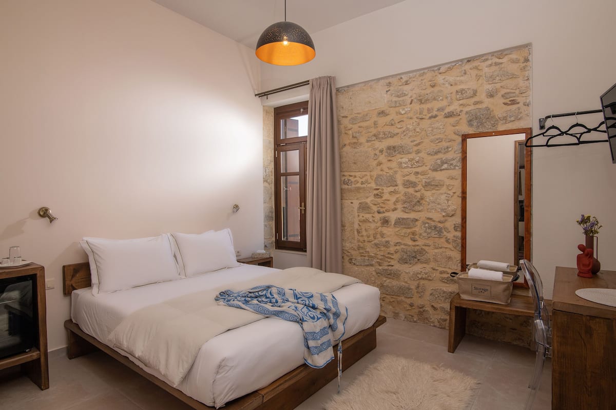 Un dormitorio con cama, mesita de noche, cómoda, espejo y pared de piedra.
