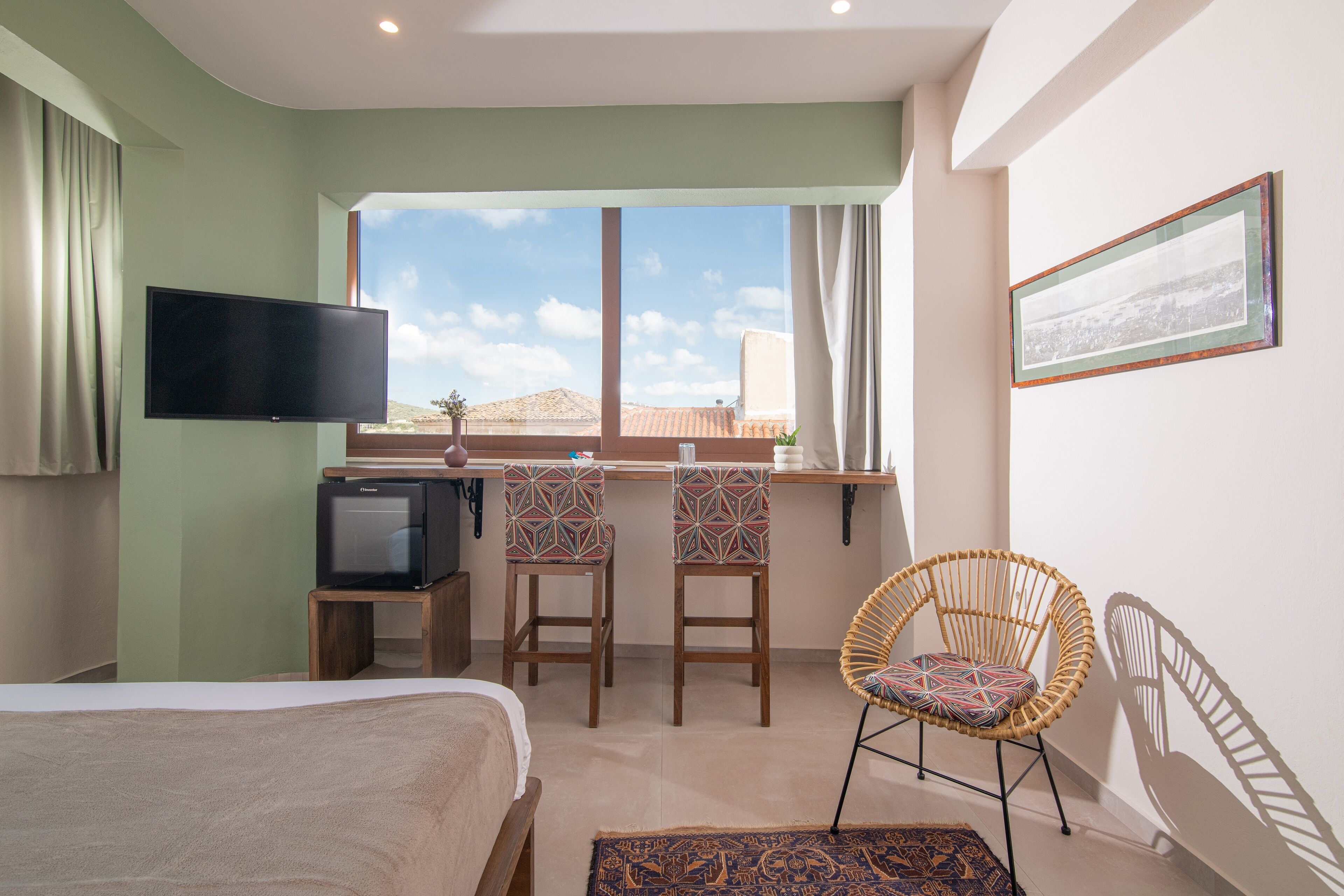A hotel room with a bed, a TV, a desk with two chairs, a wicker chair, and a window with a view of the sky and buildings.
