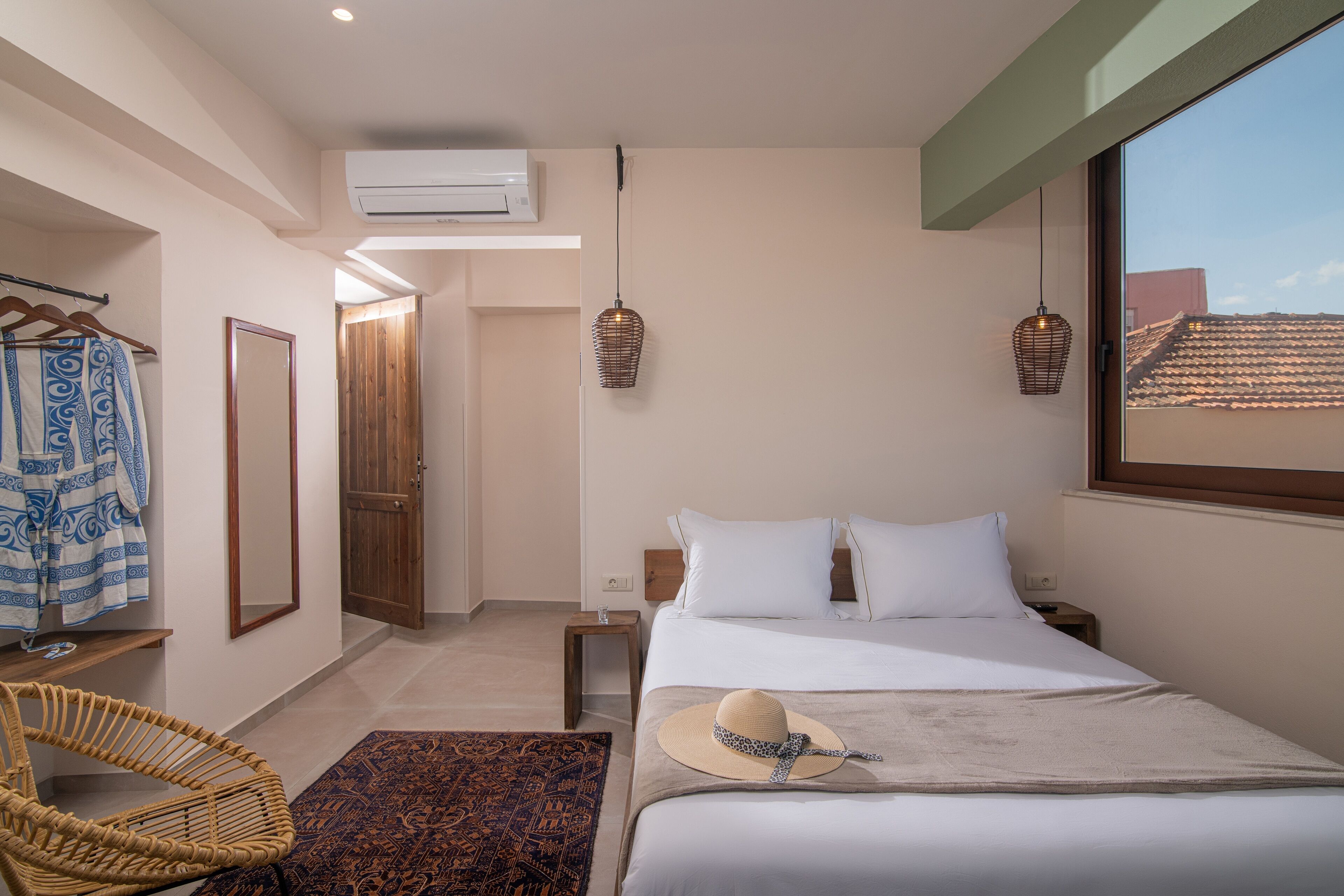 A bedroom with a bed, a hanging light fixture, a mirror, a wardrobe, and a window with a view of buildings.