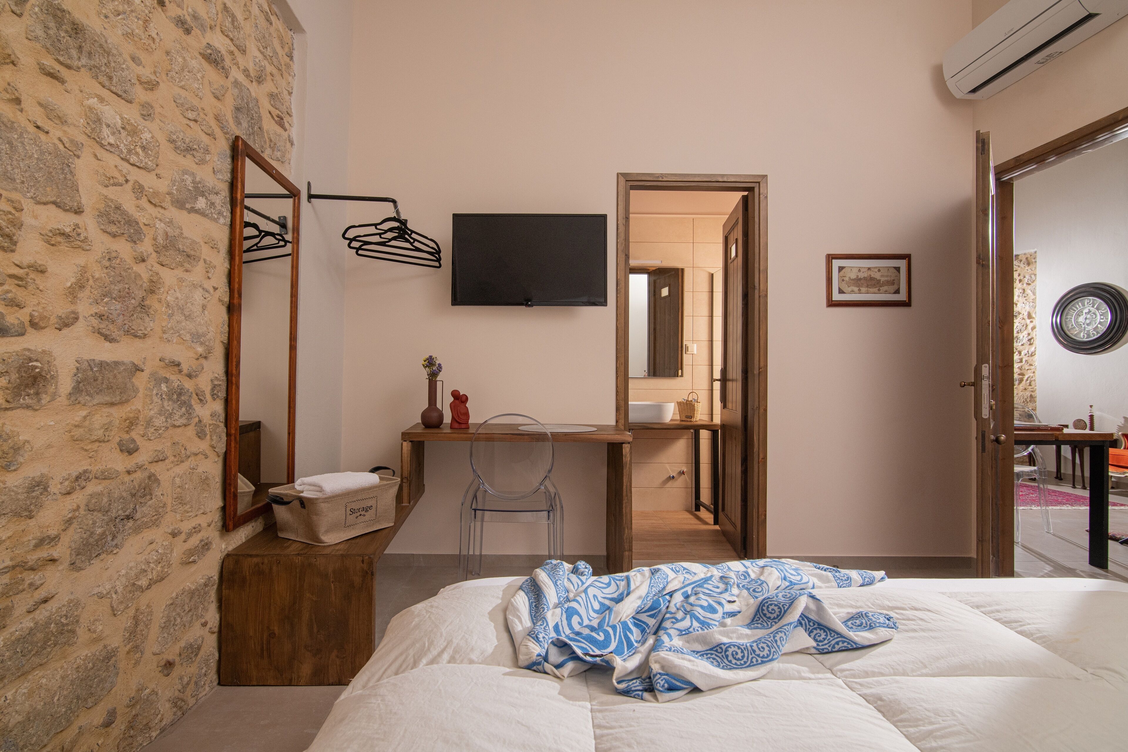 A bedroom with a stone wall, a wooden desk, a bed with a patterned blanket, a mirror, a television, and a bathroom visible through an open door.