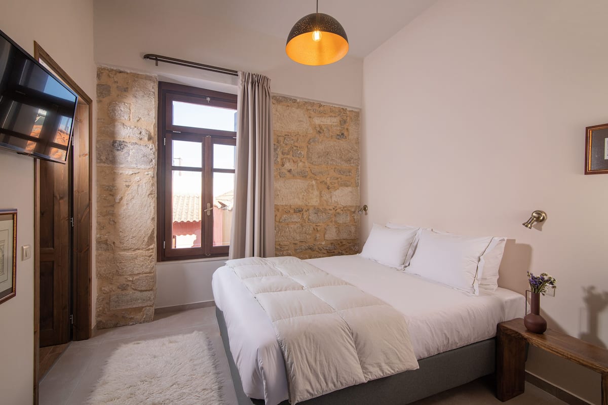 Un dormitorio con pared de piedra, cama con ropa blanca, mesita de noche de madera y lámpara colgante.