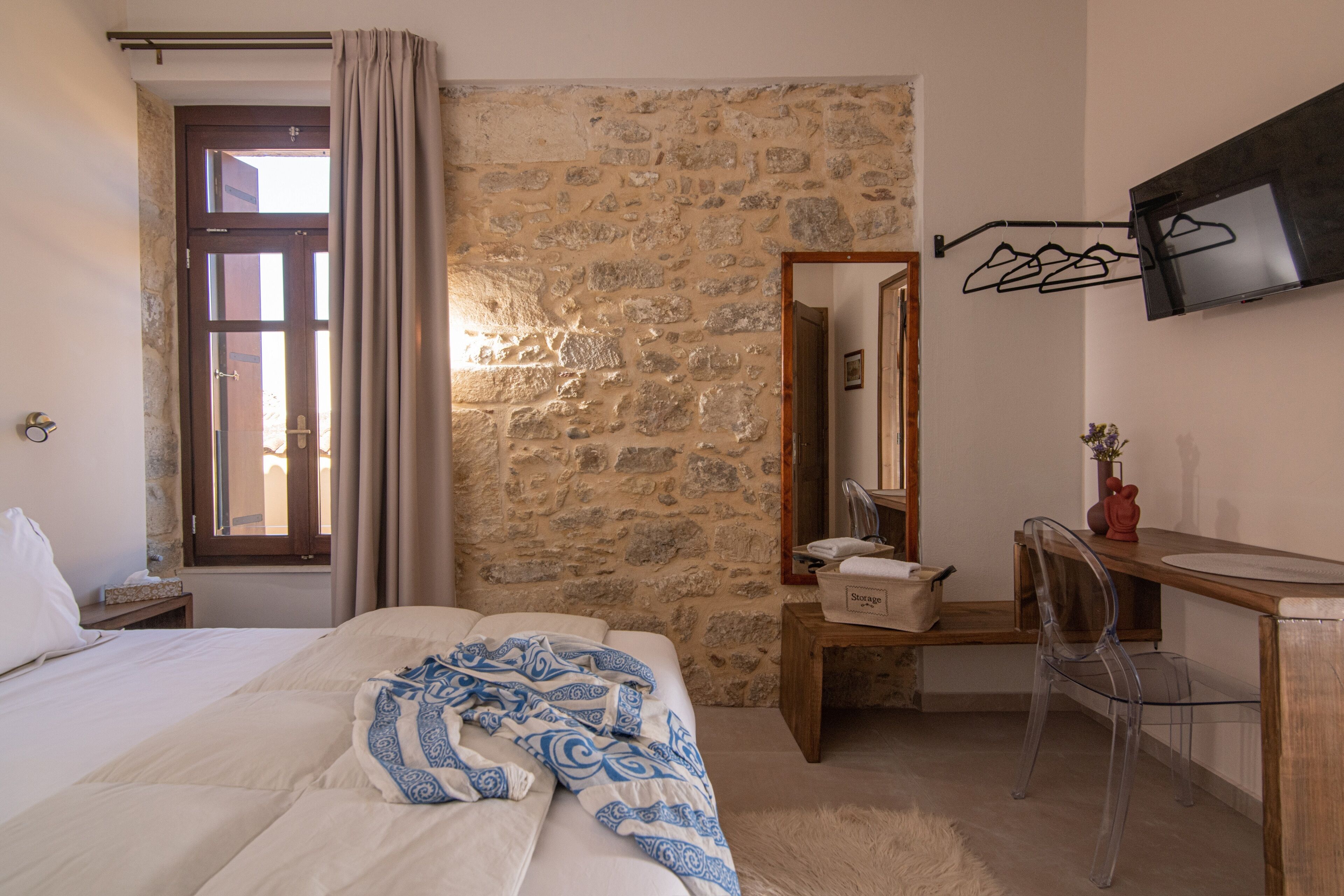 Un dormitorio con pared de piedra, cama con manta estampada, escritorio con silla, televisor montado en la pared y ventana con cortinas.
