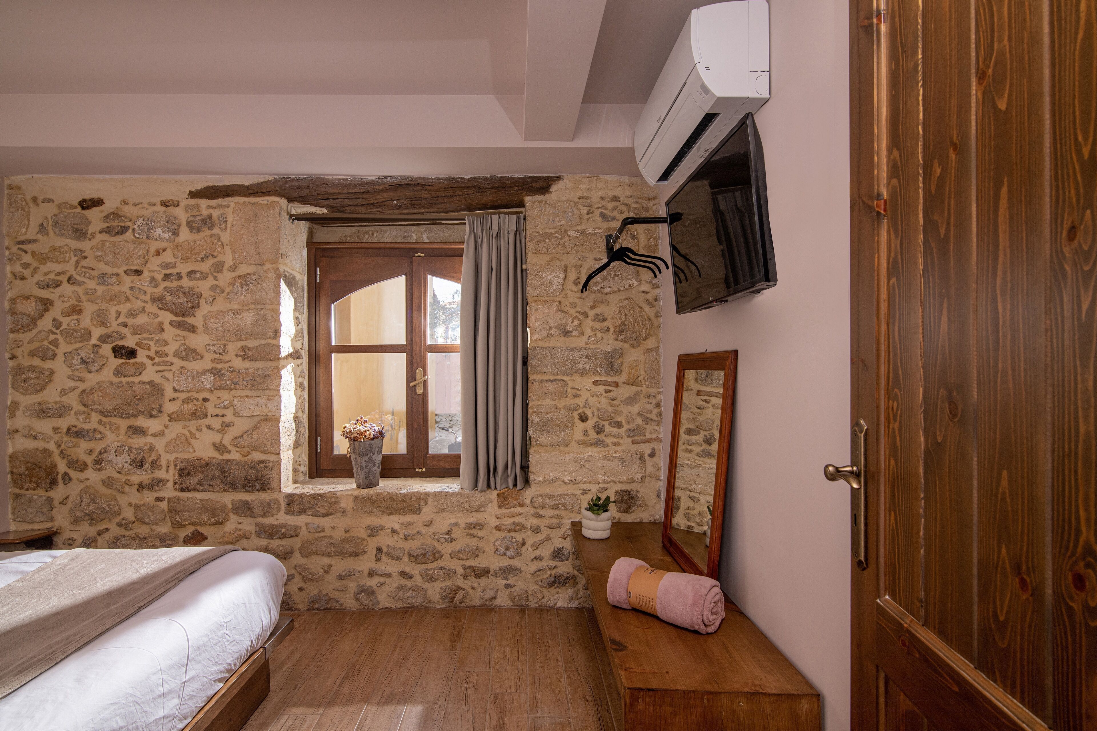 Ein Schlafzimmer mit Steinwänden, einem hölzernen Bett, einem Fenster mit Vorhängen, einer kleinen Pflanze und einer Holztür.