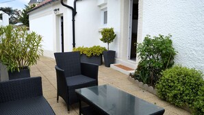 Un espacio al aire libre con mobiliario de mimbre y una mesa de vidrio, rodeado de plantas en macetas y un edificio blanco.