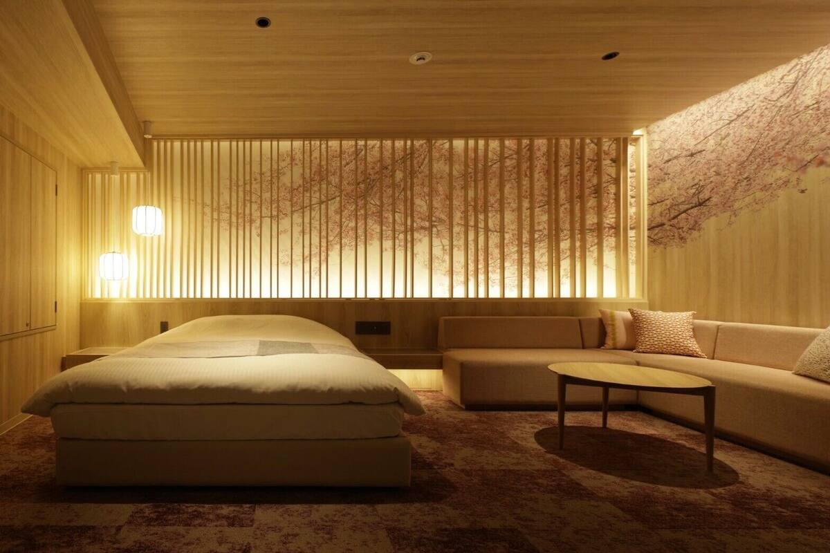 A modern hotel room with a large bed, a sofa, a small table, and a decorative wall with vertical blinds.