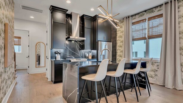 Une cuisine moderne avec des meubles de couleur sombre, un îlot central doté de tabourets de bar et une grande fenêtre équipée de stores.