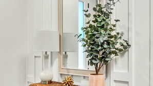 Un mur blanc avec des panneaux décoratifs, un miroir, une console en osier, une plante en pot et un vase blanc.