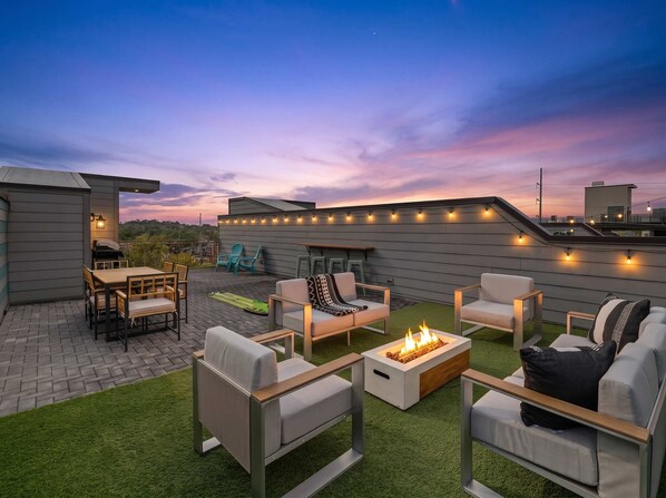 An outdoor patio area with seating, a fire feature, and string lights against a twilight sky.