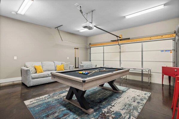 A pool table in a room with a grey sofa, a red cabinet, and a garage door.