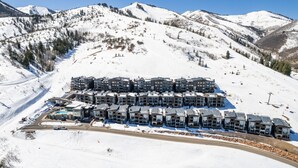 A snow-covered mountain area with a row of multi-story buildings, likely a hotel, with a ski slope in the background. - Apex Ski In/Out Canyons Village Beautiful 3 Bedroom, Private Hot Tub, Community Pool (Park City)