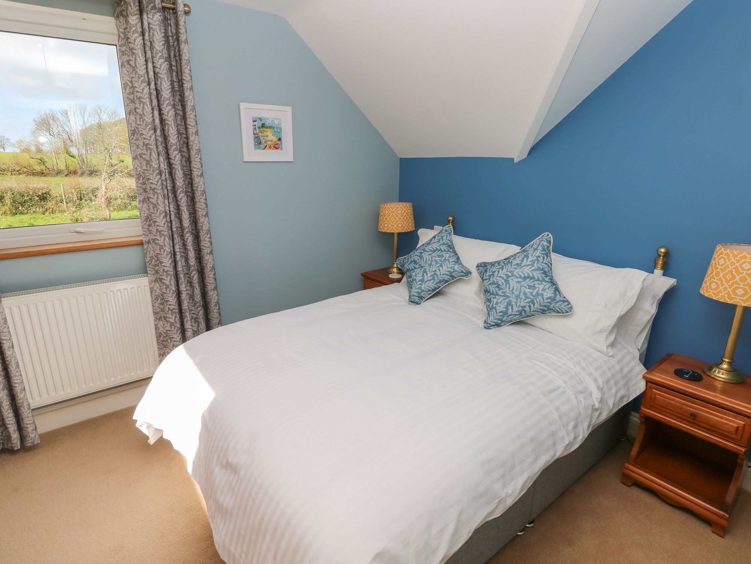 A bedroom with a large bed, two bedside tables with lamps, a window with curtains, and a radiator.