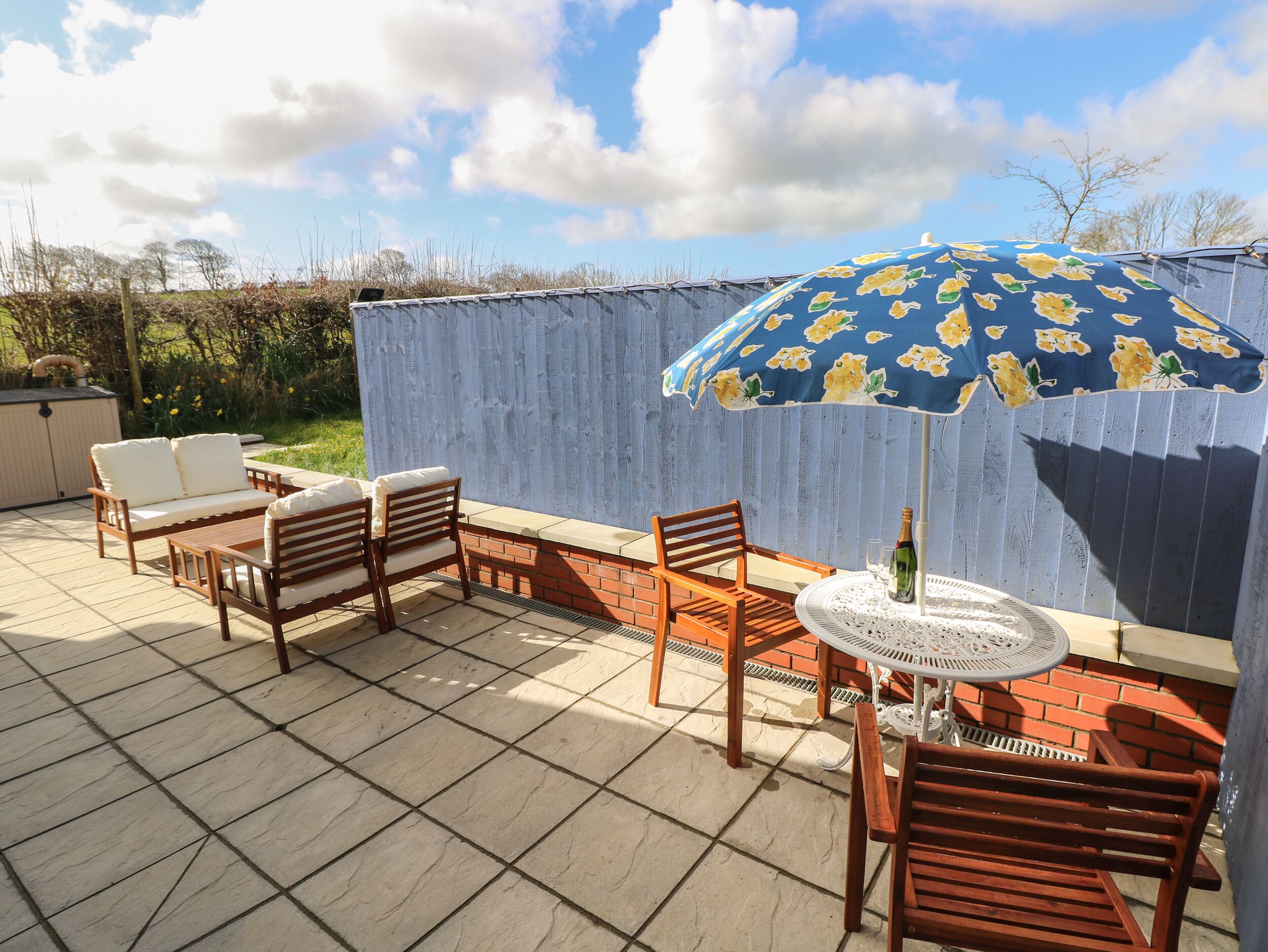 An outdoor patio with a wooden table and chairs, a blue floral umbrella, and a white decorative table base.