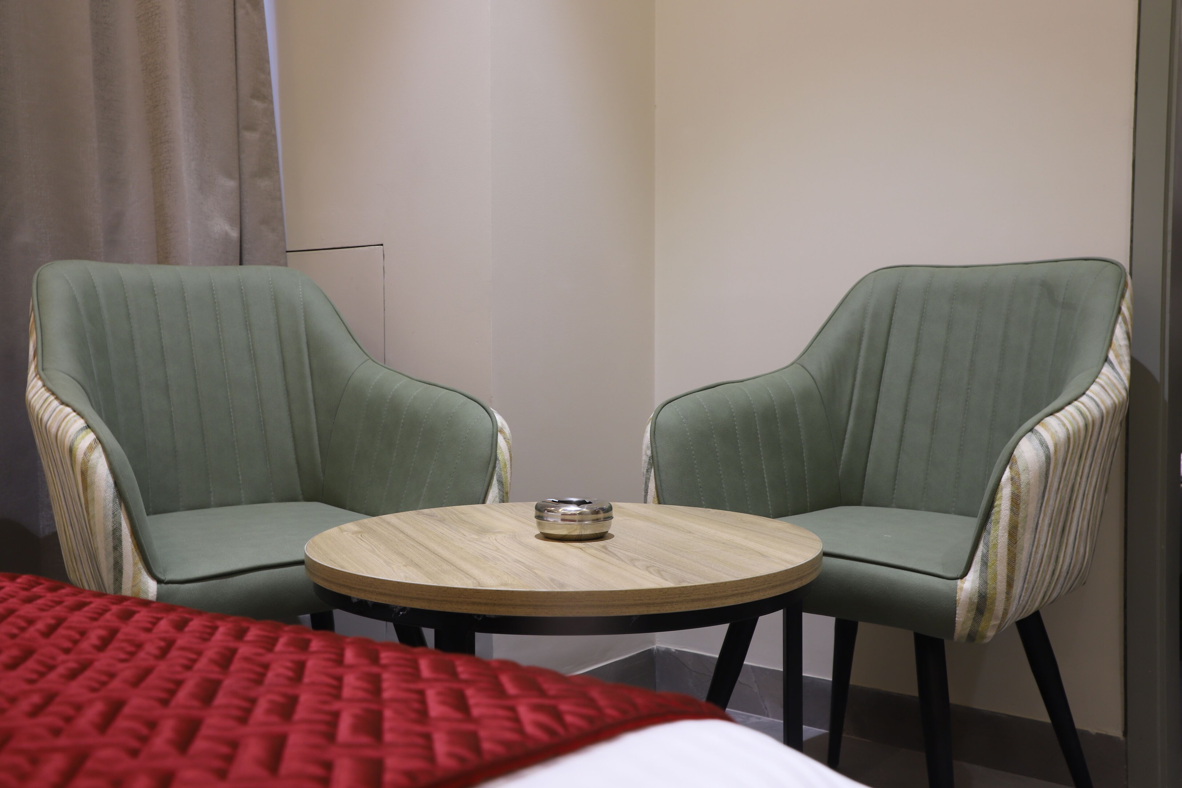 Habitación de hotel con dos sillones tapizados en verde alrededor de una pequeña mesa de madera, una colcha de patchwork roja y una pared lisa con cortina.