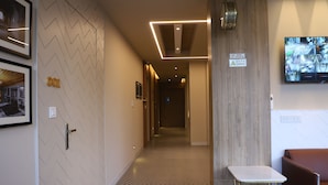Un couloir d’hôtel avec un sol à motifs en chevron, un comptoir d’accueil, un espace détente et un panneau indiquant la présence de caméras de surveillance.