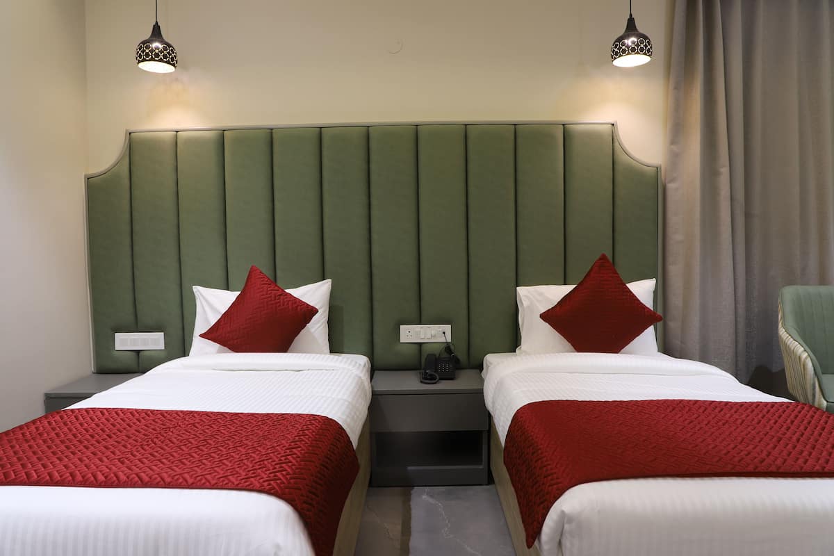 A hotel room with two beds, green headboard, red and white bedding, bedside tables with lamps, and a chair.
