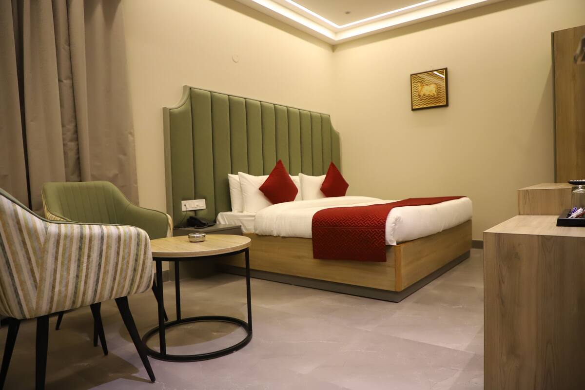 A hotel room with a bed, a green upholstered chair, a small round table, and a wooden desk.