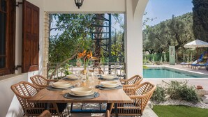 Uma área de refeições com mesa posta para quatro pessoas, cercada por cadeiras de vime, um candeeiro pendente e vista para uma piscina e um jardim.