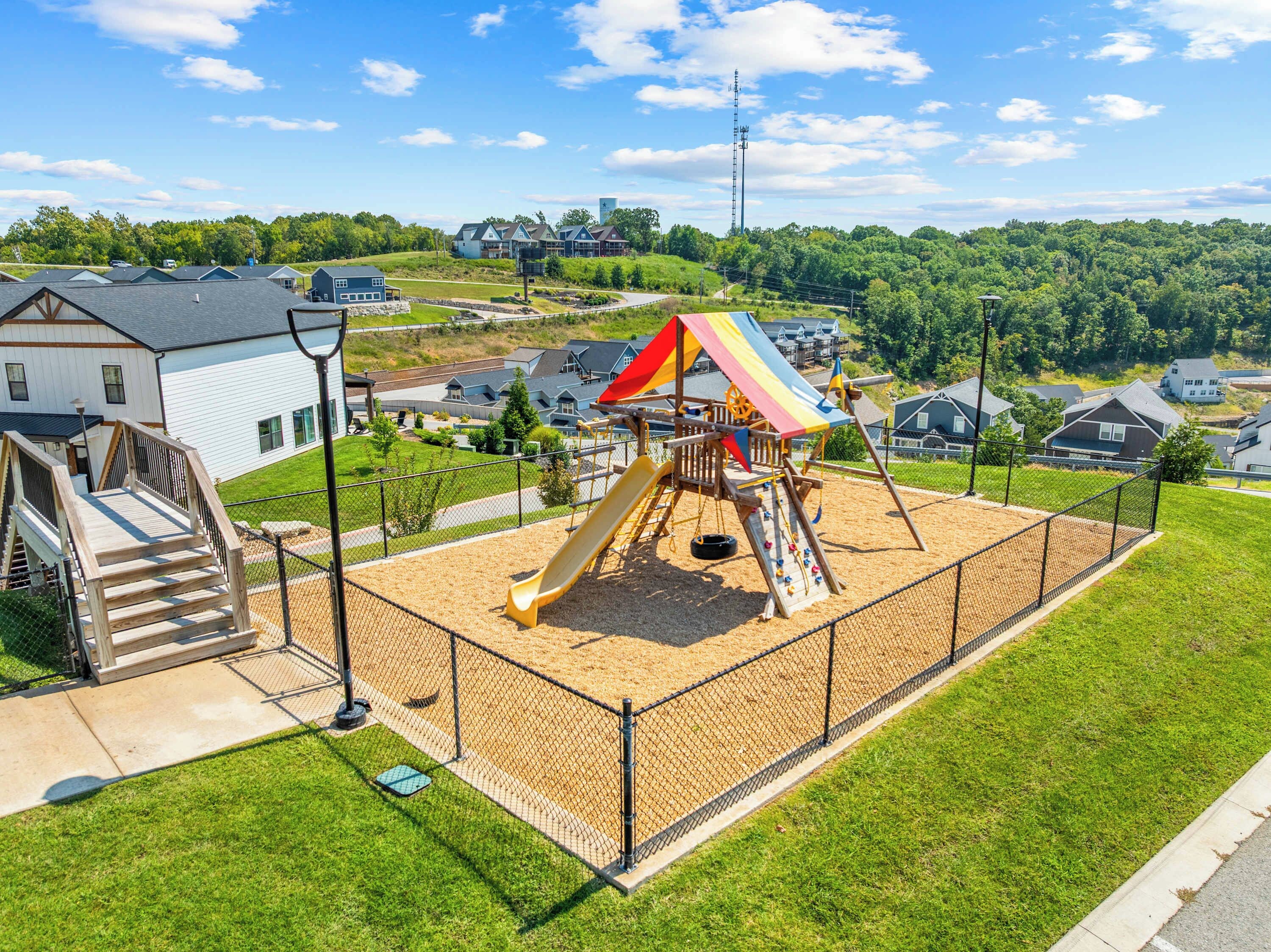 A playground with a slide and climbing structures, surrounded by a fence, with houses in the background.