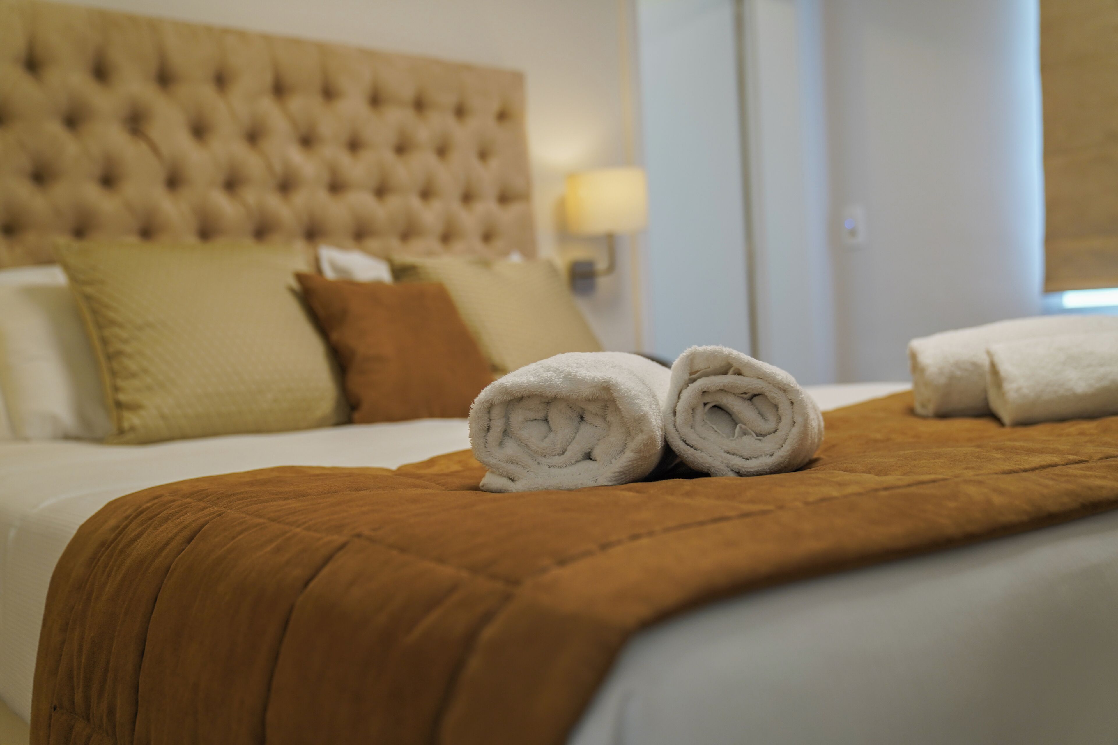 A neatly made bed with a brown blanket and rolled white towels, a tufted headboard, and a bedside lamp.