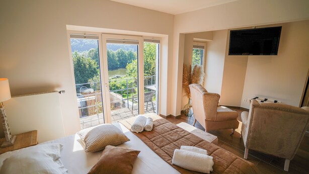 Habitación de hotel con cama, una silla, un televisor y un balcón con vistas a árboles y un lago.