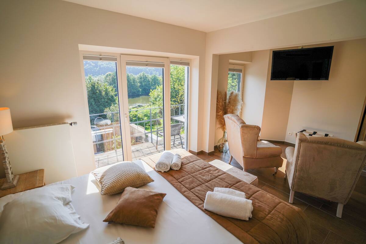 Ein Hotelzimmer mit Bett, Sessel, Fernseher und einem Balkon mit Blick auf Bäume und See.