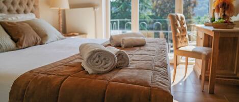 Una cama bien tendida con un edredón marrón, una toalla enrollada y una silla con una mesita de madera al lado.