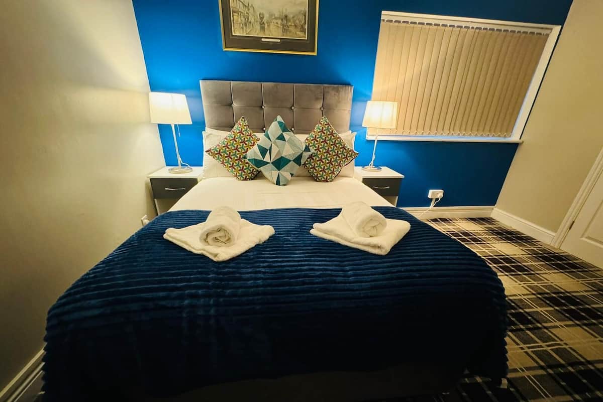 Habitación de hotel con pared azul, cabecero capitonado, piso estampado de cuadros y dos toallas sobre la cama.