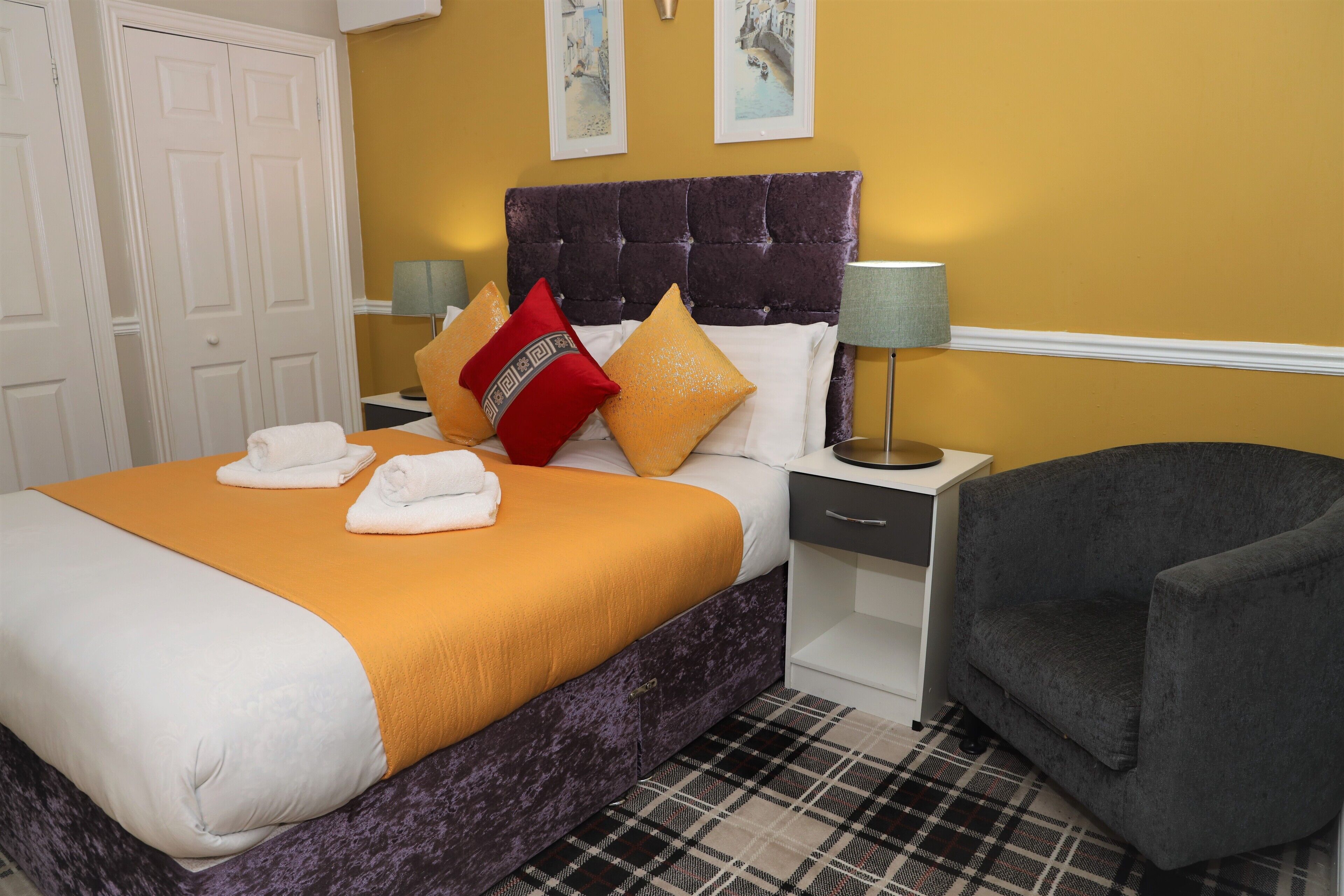Una cama bien tendida con un cabecero morado, un tapete escocés y un sillón gris.