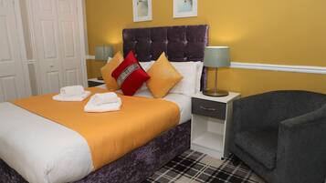 Una cama bien tendida con un cabecero morado, un tapete escocés y un sillón gris.