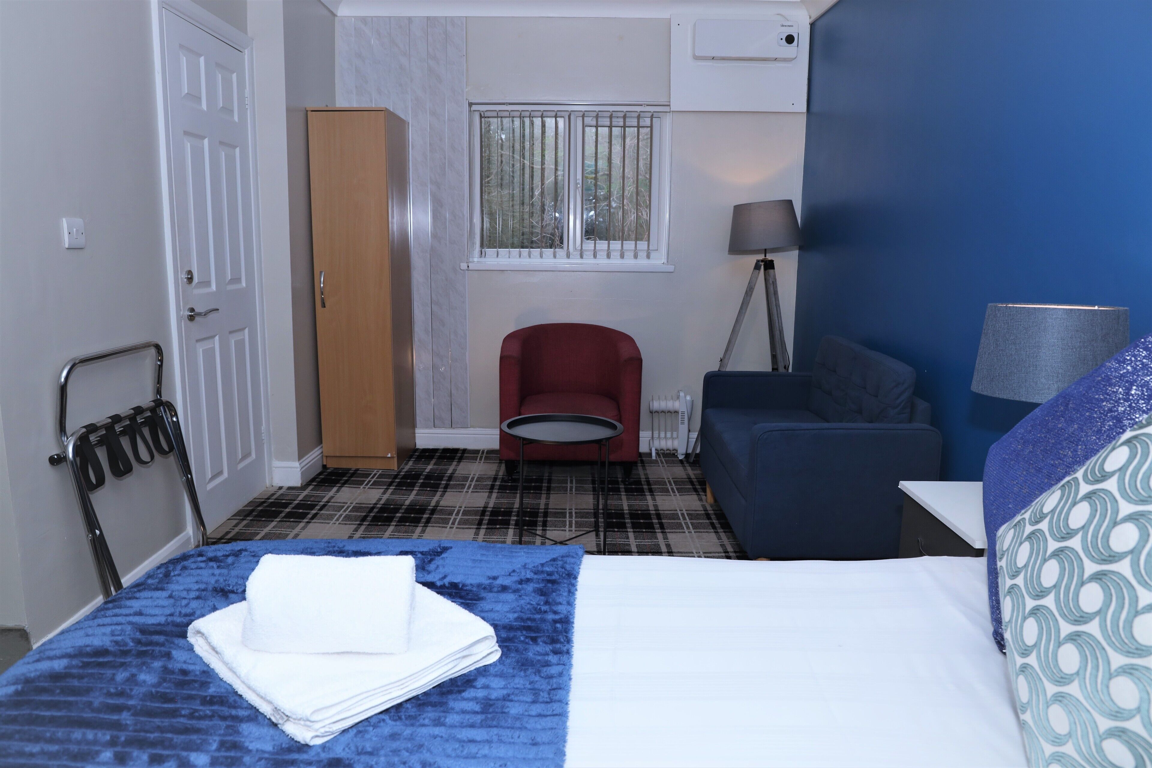A hotel room with a bed, a red chair, a blue sofa, a lamp, and a window with bars.