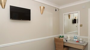 Un baño de hotel con televisor de pantalla plana, un espejo grande, un lavamanos con artículos de aseo y una silla de mimbre.