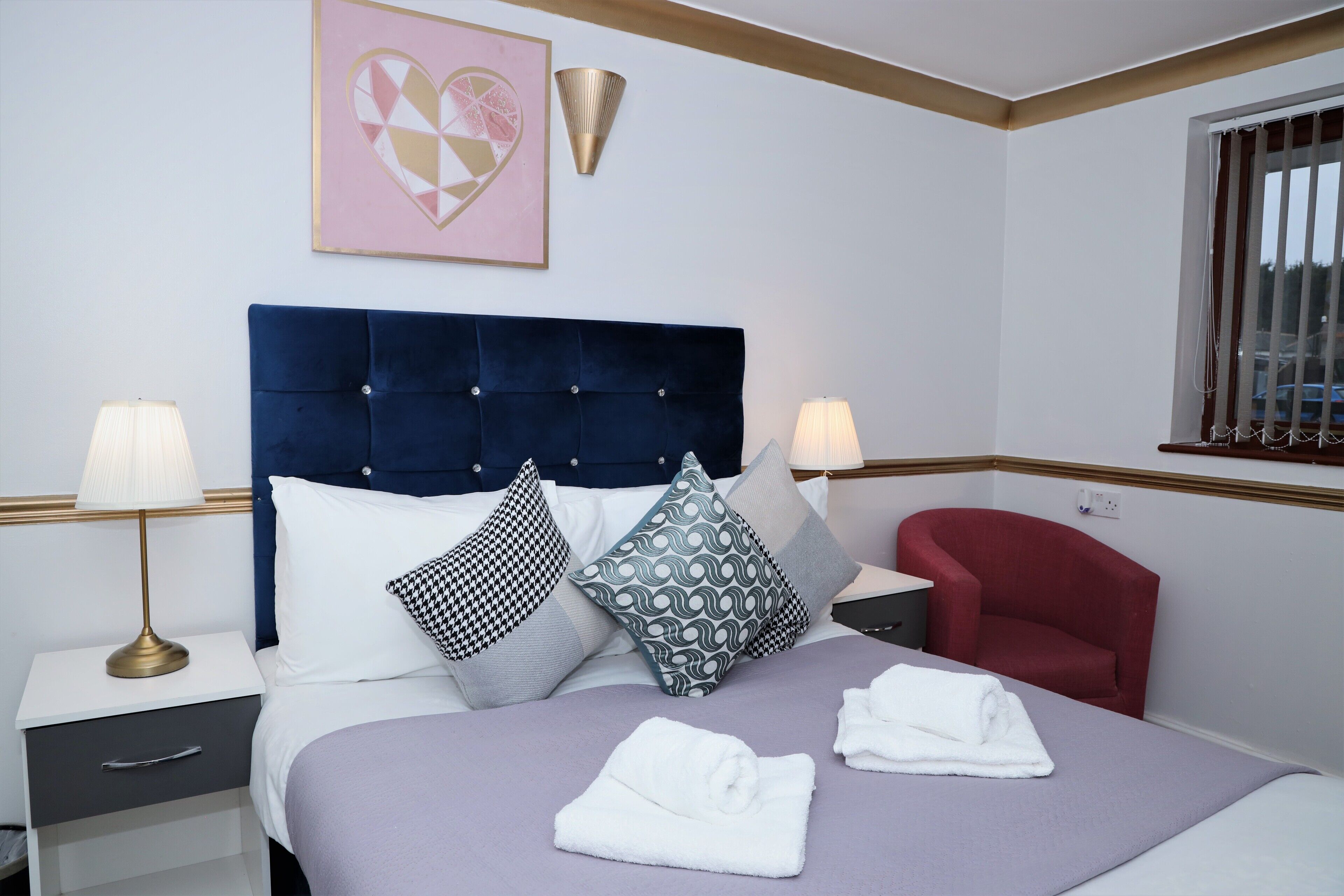 A neatly arranged hotel room with a large bed, two bedside tables, a red armchair, and a window with blinds.
