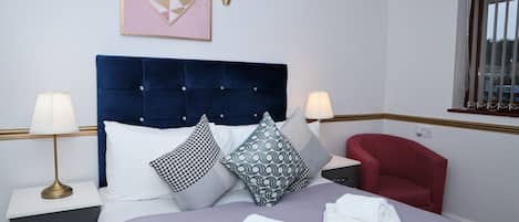 Une chambre d’hôtel bien aménagée, avec un grand lit, deux tables de chevet, un fauteuil rouge et une fenêtre avec des stores.