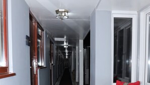 Un pasillo largo y estrecho con alfombra oscura, paredes blancas y una silla de mimbre con cojines rojos y amarillos.
