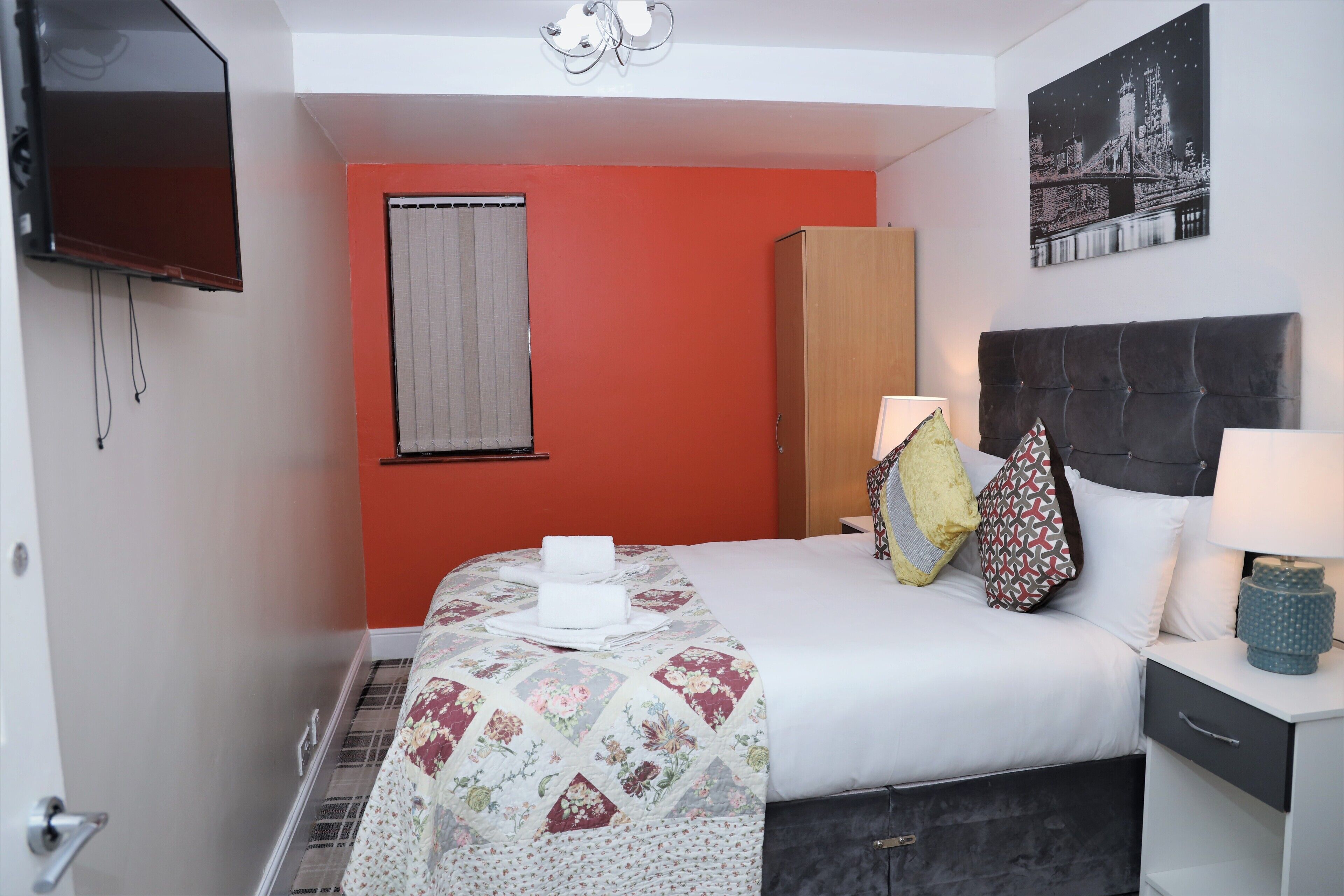 A hotel room with a bed, a nightstand, a TV, a window with blinds, and a cityscape painting on the wall.