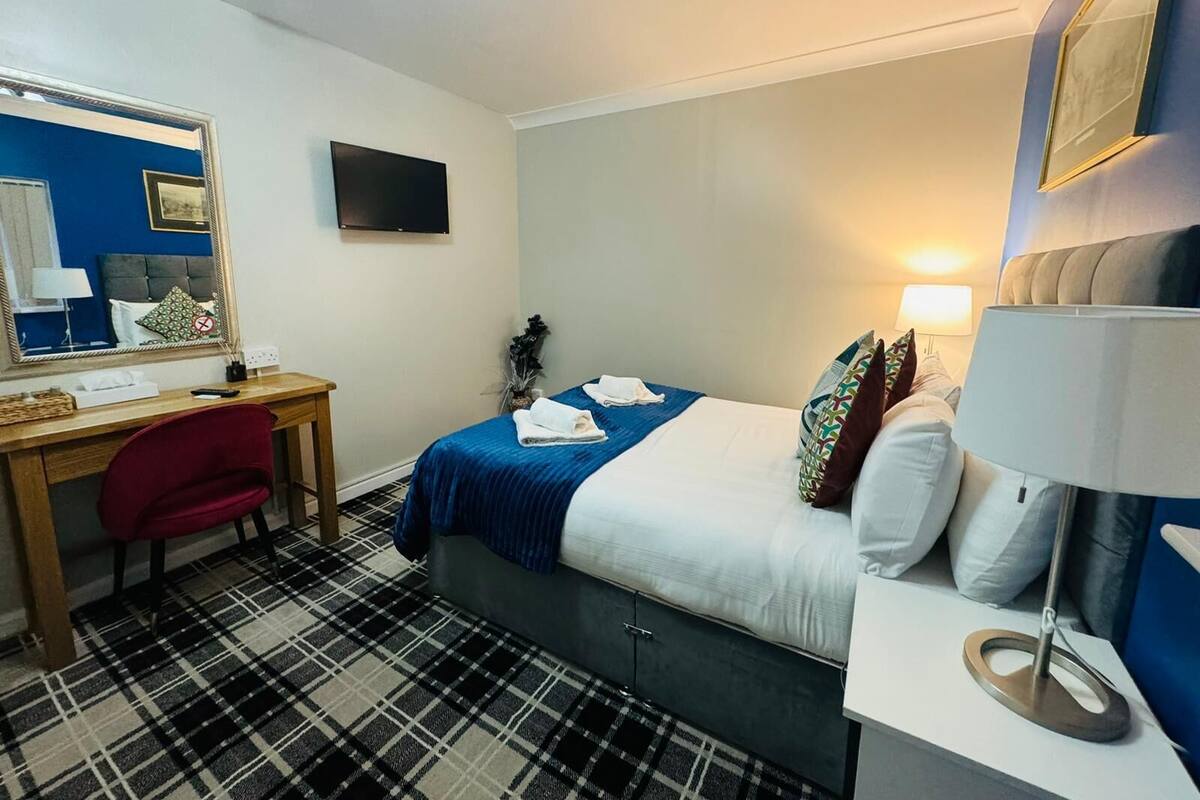 Habitación de hotel con cama, escritorio, silla, espejo, televisor y lámpara.
