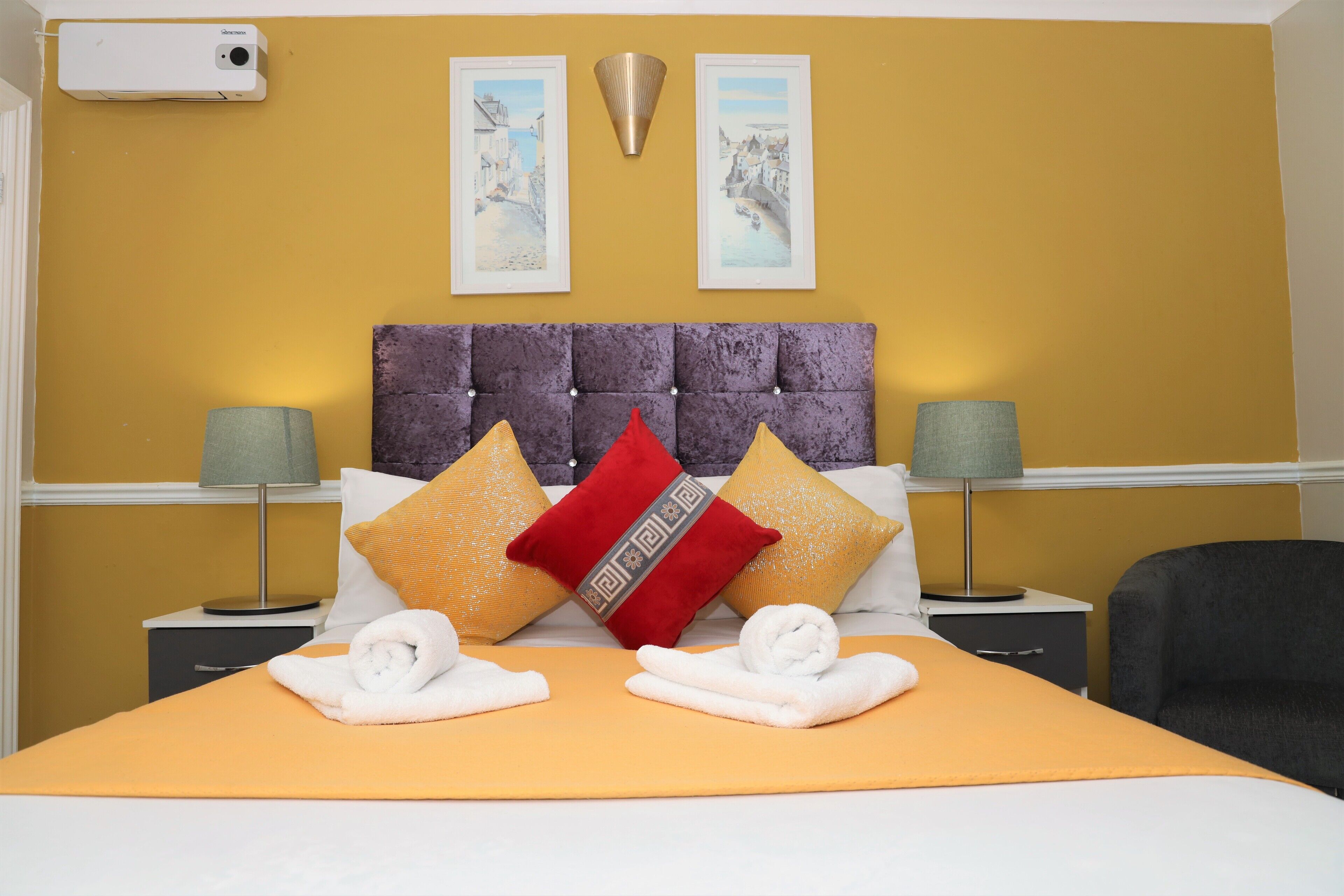 A hotel room with a yellow wall, a tufted headboard, two bedside tables with lamps, and a bed with towels and decorative pillows.