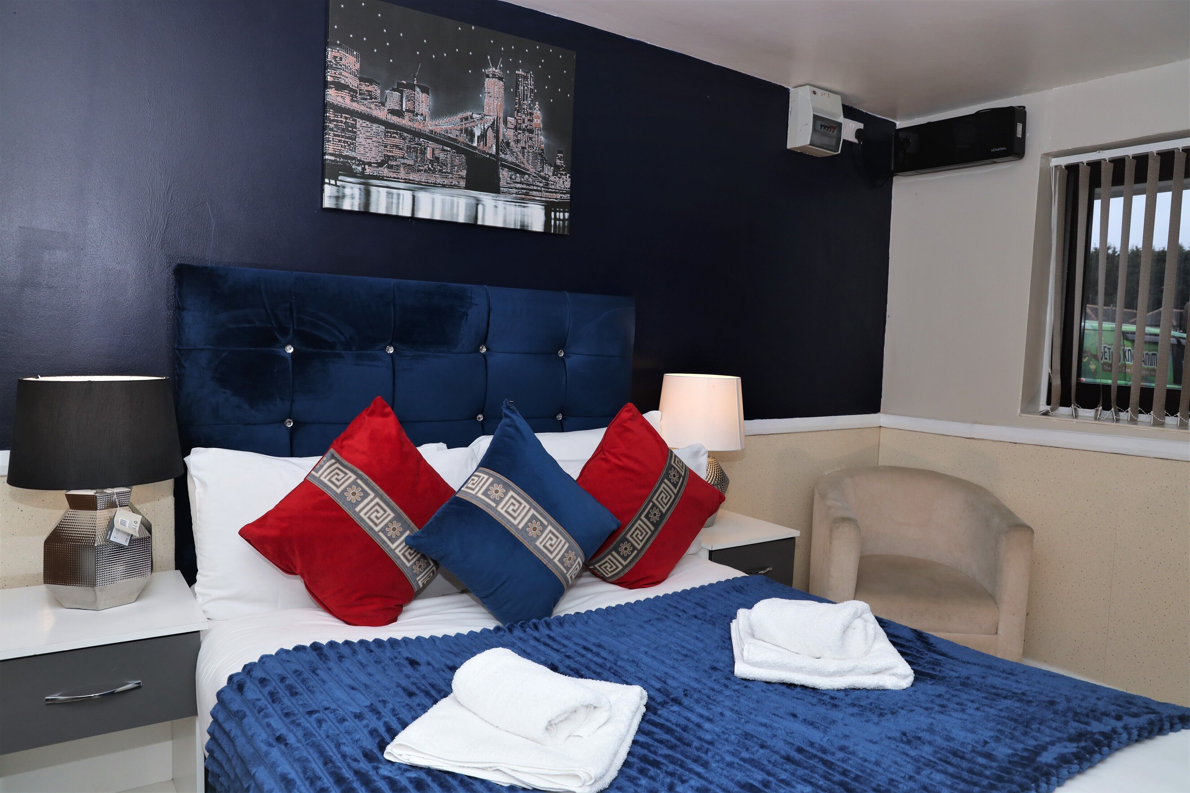 A hotel room with a large bed, a nightstand, a chair, a window with bars, and a cityscape painting on the wall.
