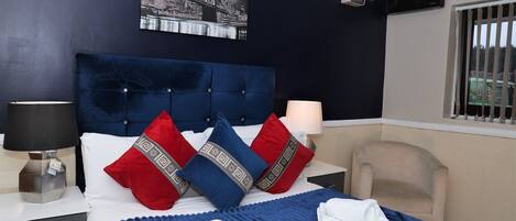 A hotel room with a large bed, a nightstand, a chair, a window with bars, and a cityscape painting on the wall.