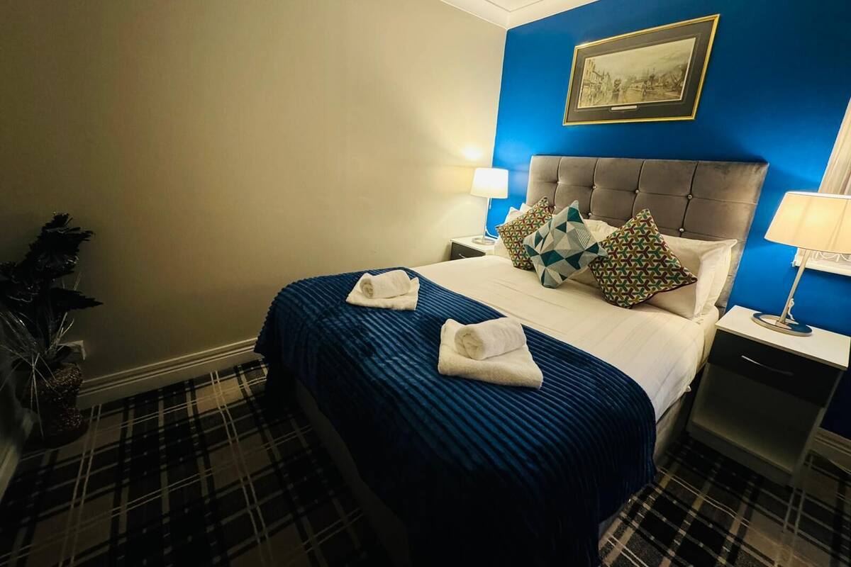 Una habitación de hotel con una cama grande, cabecera, mesita de noche, lámpara, un cuadro en la pared y una planta.