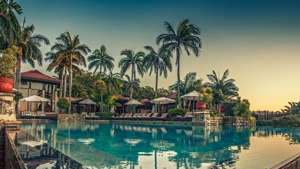 Zwembad van een resort met ligbedden en parasols, omgeven door palmbomen en een gebouw met een rood dak.