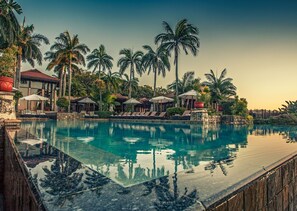 Zwembad van een resort met ligbedden en parasols, omgeven door palmbomen en een gebouw met een rood dak.