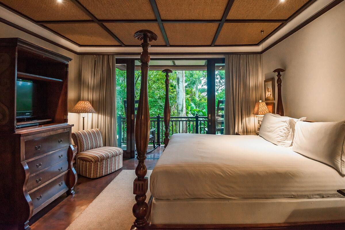 Une chambre spacieuse avec un grand lit, une télévision, une commode en bois et un balcon offrant une vue sur la verdure.