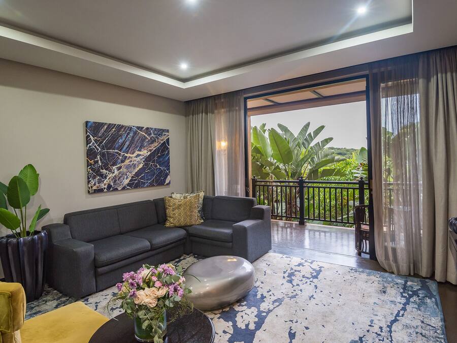 A modern living room with a grey sofa, a coffee table, and a vase of flowers. There is a large painting on the wall and a balcony with plants.