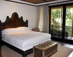 Una cama grande con cabecera de madera, una mesita y vistas a un balcón con plantas.