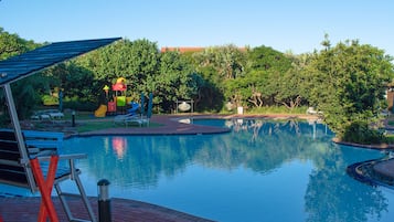 Relax by the serene pool surrounded by lush greenery and trees, with lounge chairs and a shaded area for unwinding.