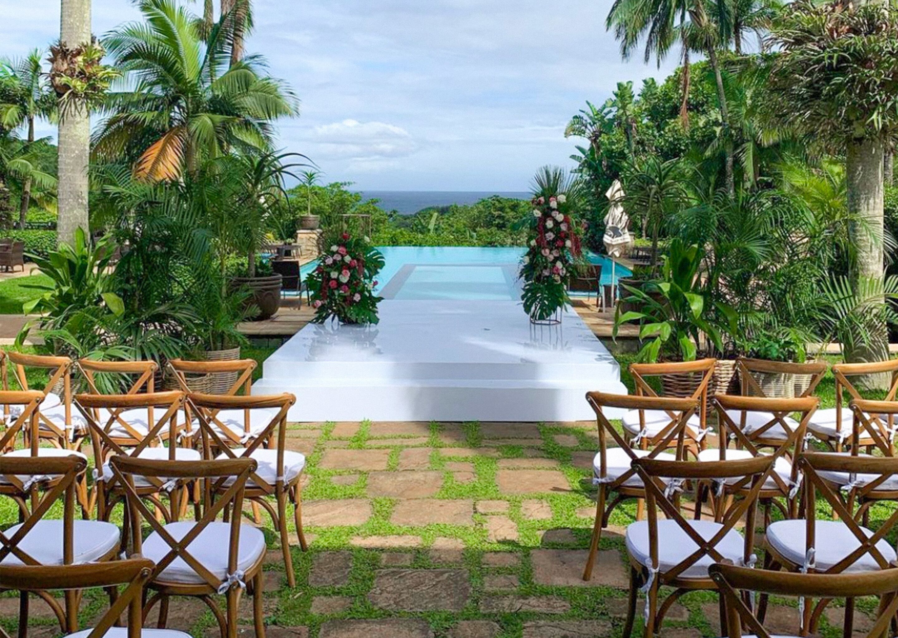 Pasillo nupcial al aire libre con sillas y decoración floral, que conduce a una piscina.